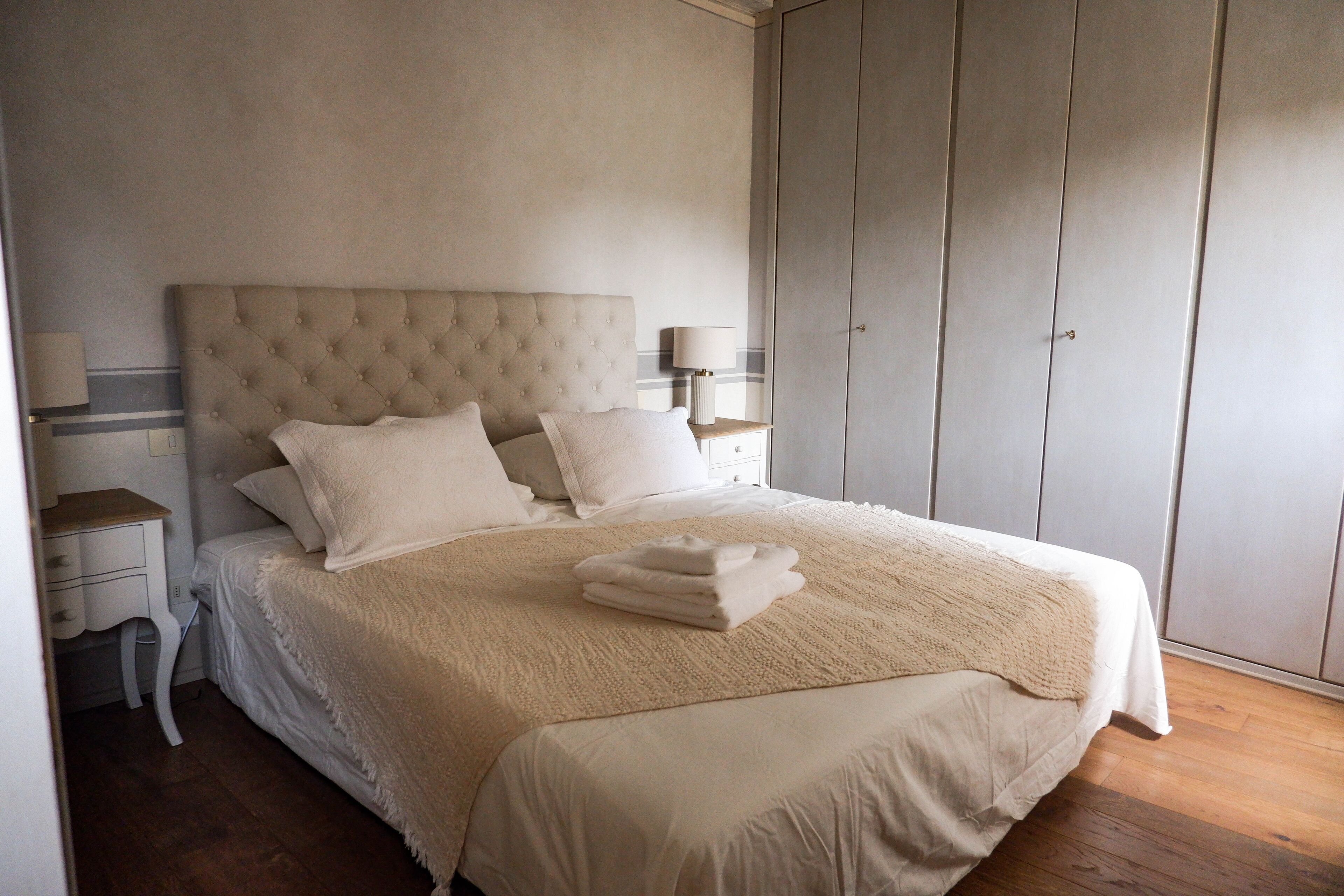 Une chambre à coucher avec une tête de lit capitonnée, un grand lit avec du linge de lit blanc, une table de chevet avec une lampe et une armoire.