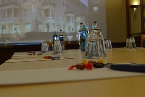 Una mesa de conferencias con cristalería transparente, una pantalla de proyector mostrando una imagen de un edificio y una vista parcial del interior de un hotel.