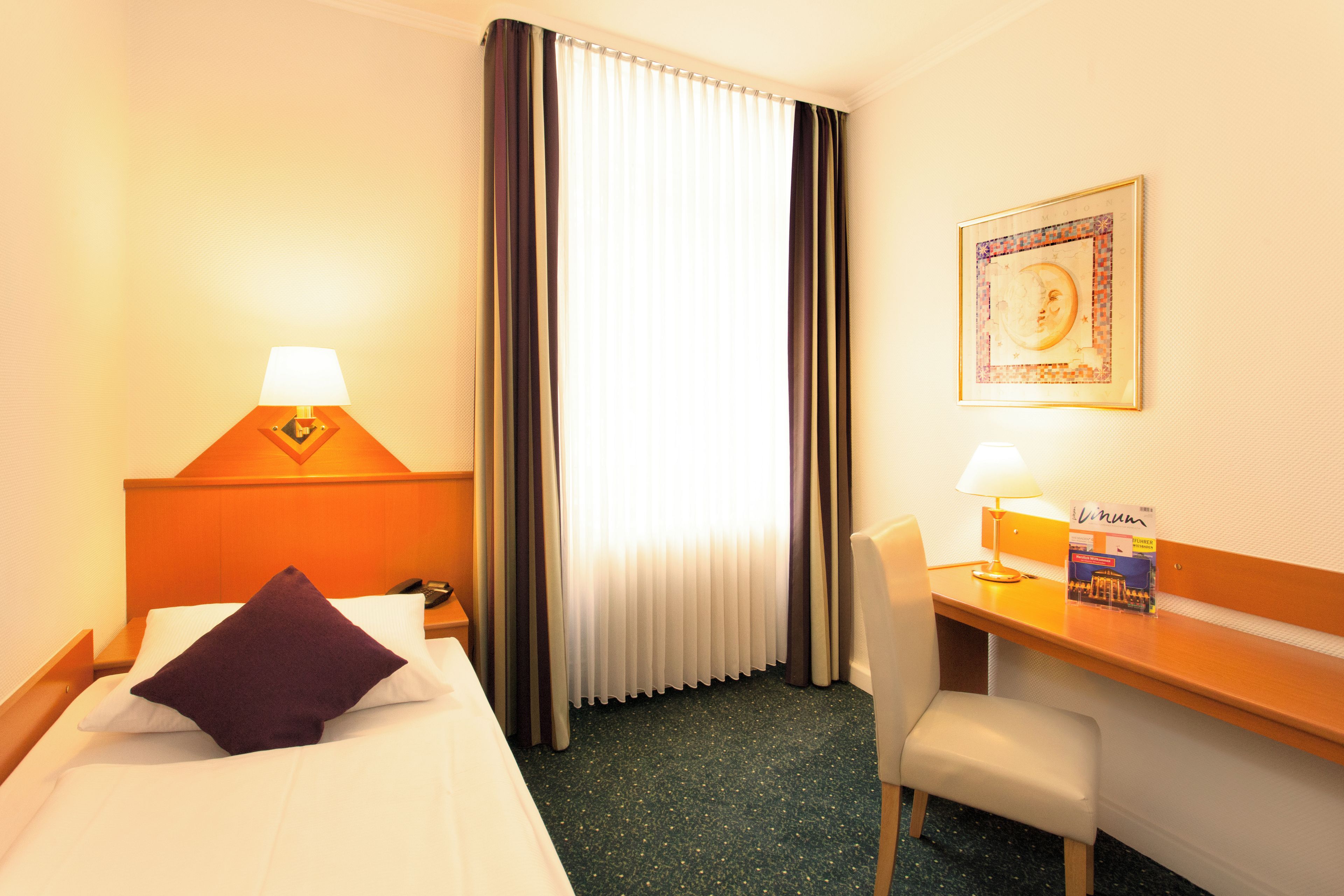 A hotel room with a bed, a desk, a chair, a lamp, a telephone, and a framed picture on the wall.