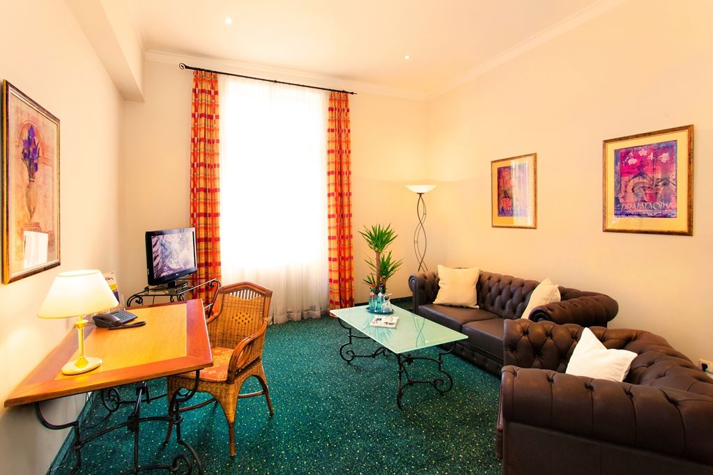 A hotel room with a television, a desk with a lamp, a sofa, a coffee table, and a large window with curtains.
