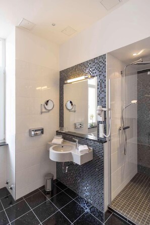 A modern bathroom with a black and white tiled wall, a round mirror, a sink, and a shower area. - TOP Hotel Hansa Wiesbaden (Wiesbaden)