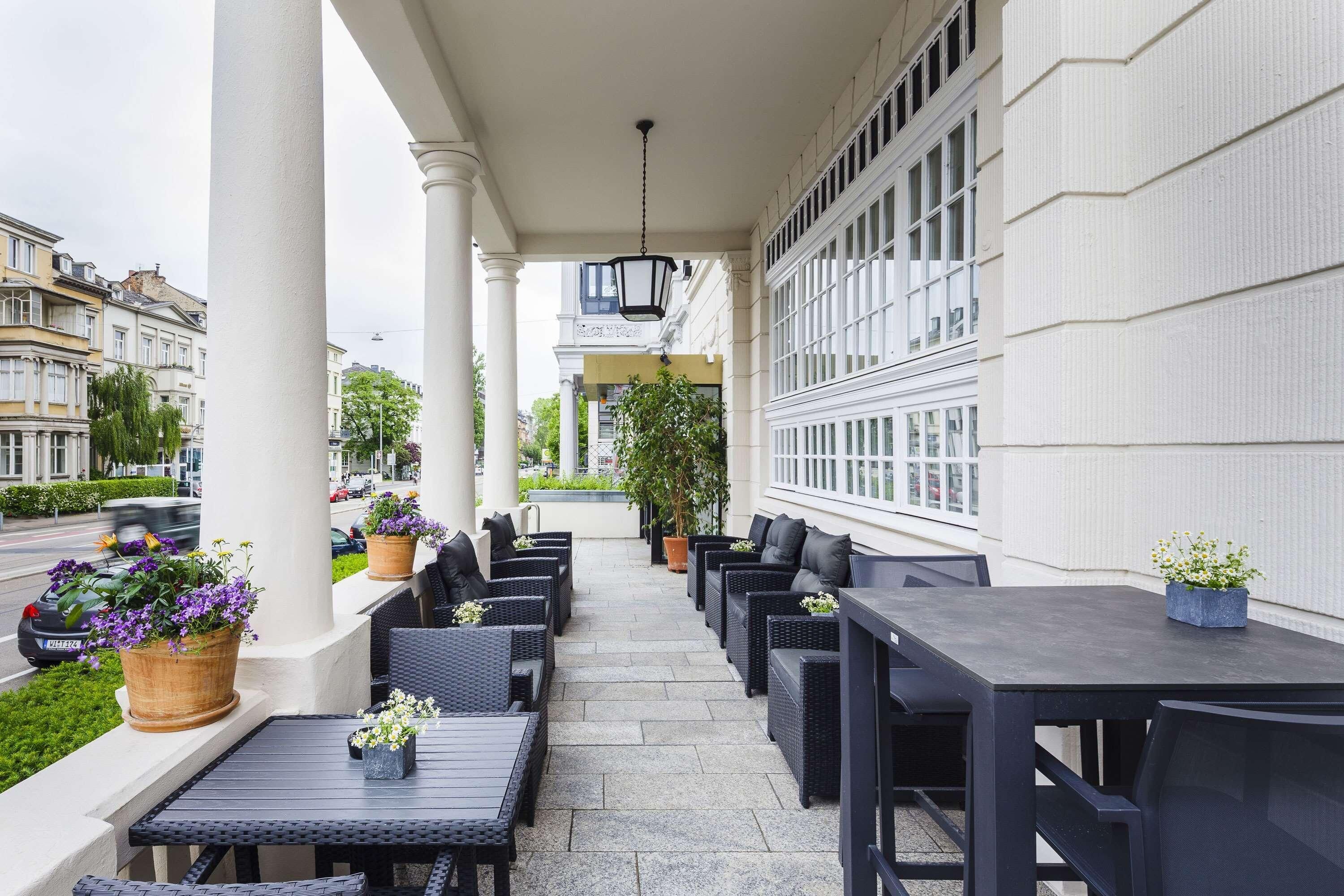 An outdoor seating area with tables and chairs, potted plants, and a view of the street.