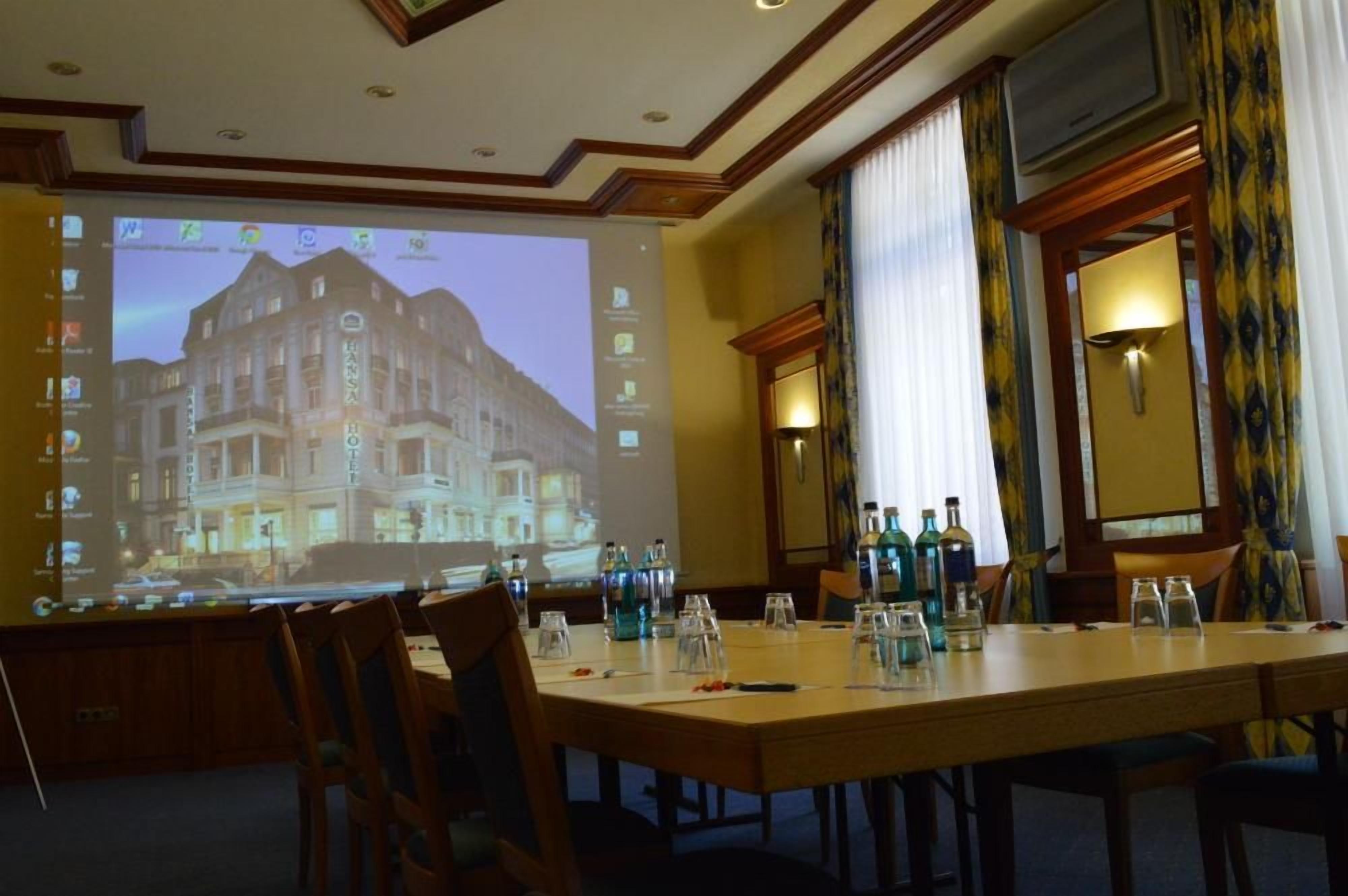 Ein Konferenzraum mit einer Projektionsleinwand, die ein Hotel zeigt, einem langen Tisch mit Stühlen und einem Fenster mit Vorhängen.
