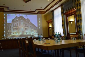 Une salle de conférence équipée d’un écran de projection affichant une image d’hôtel, d’une longue table entourée de chaises et d’une fenêtre avec des rideaux.