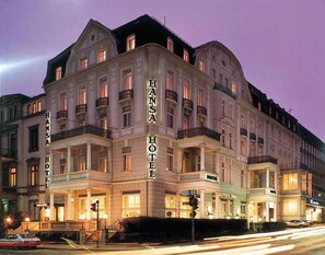 Um edifício histórico com o nome "Hansa Hotel" em destaque. Apresenta um estilo arquitetônico clássico, com várias varandas e janelas grandes.