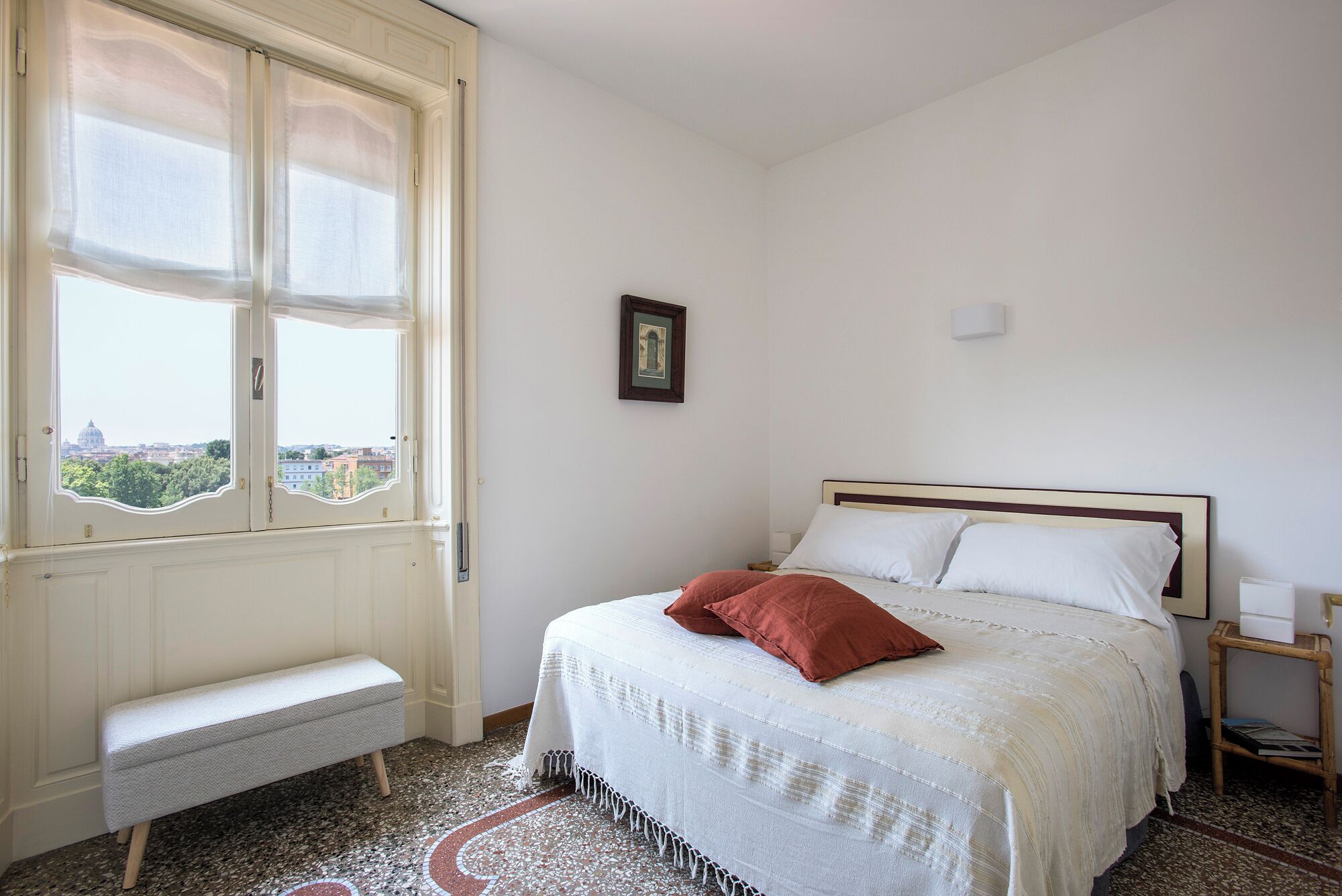 A bedroom with a bed, a bench, a framed picture, and a view of a cityscape through a window.