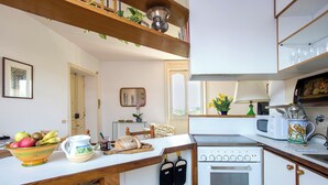 Une cuisine compacte équipée d’une cuisinière, d’un four à micro-ondes et d’un petit four. On y trouve un comptoir en bois sur lequel est posé un saladier rempli de fruits et une planche à découper. Une chaise est visible au fond.