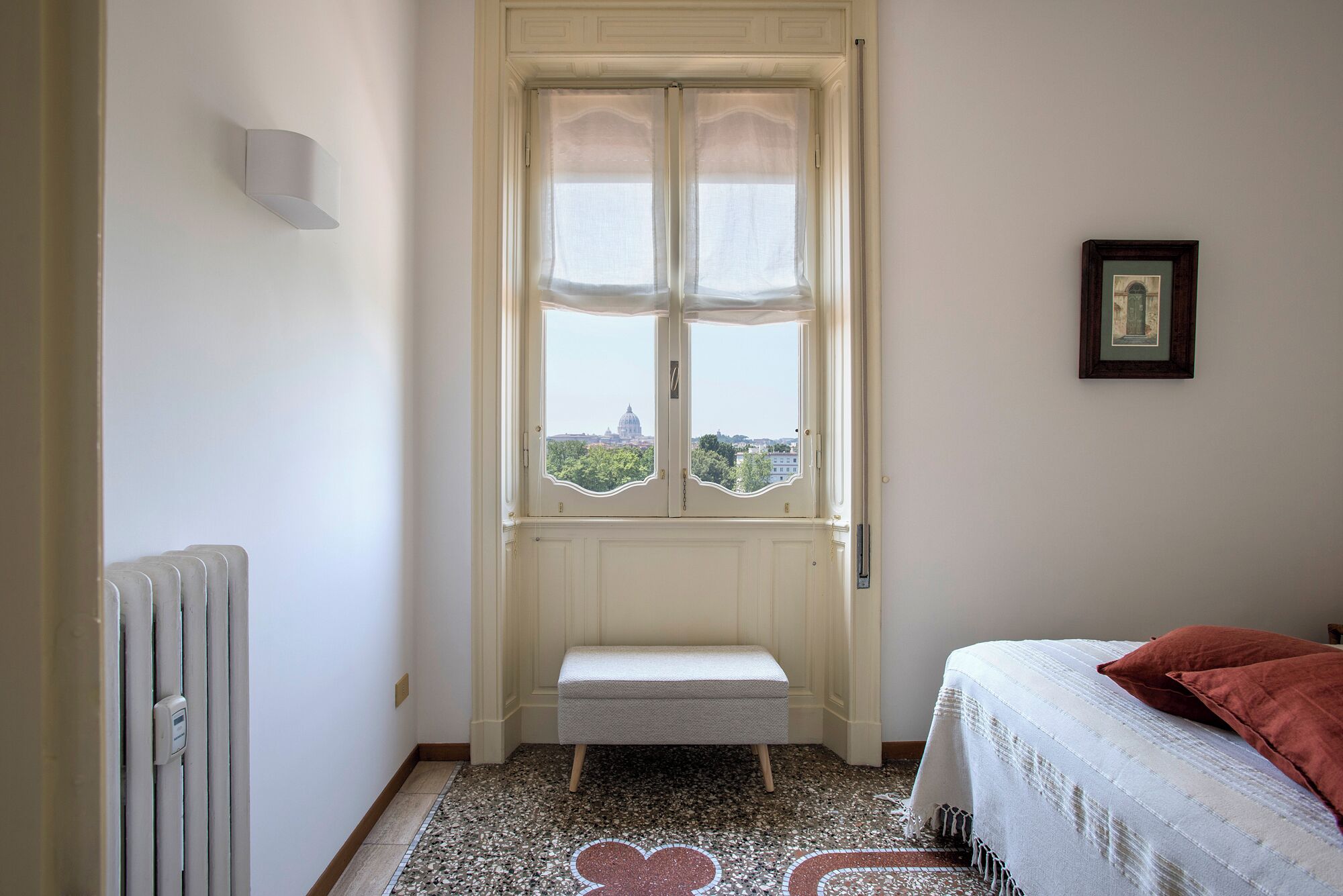 Un dormitori amb llit, una banca, un radiador, una finestra amb vista i un quadre amb marc a la paret.