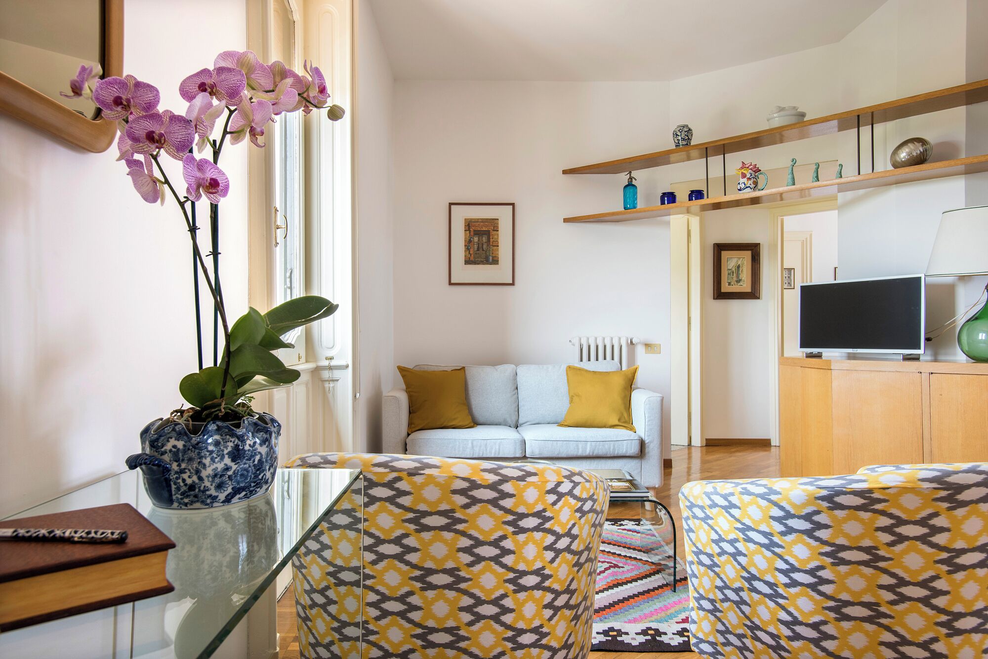 A living room with a sofa, patterned armchairs, a coffee table, a TV, and decorative items on shelves.