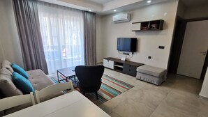 A modern living room with a sofa, a dining table, a TV, and a small kitchenette.