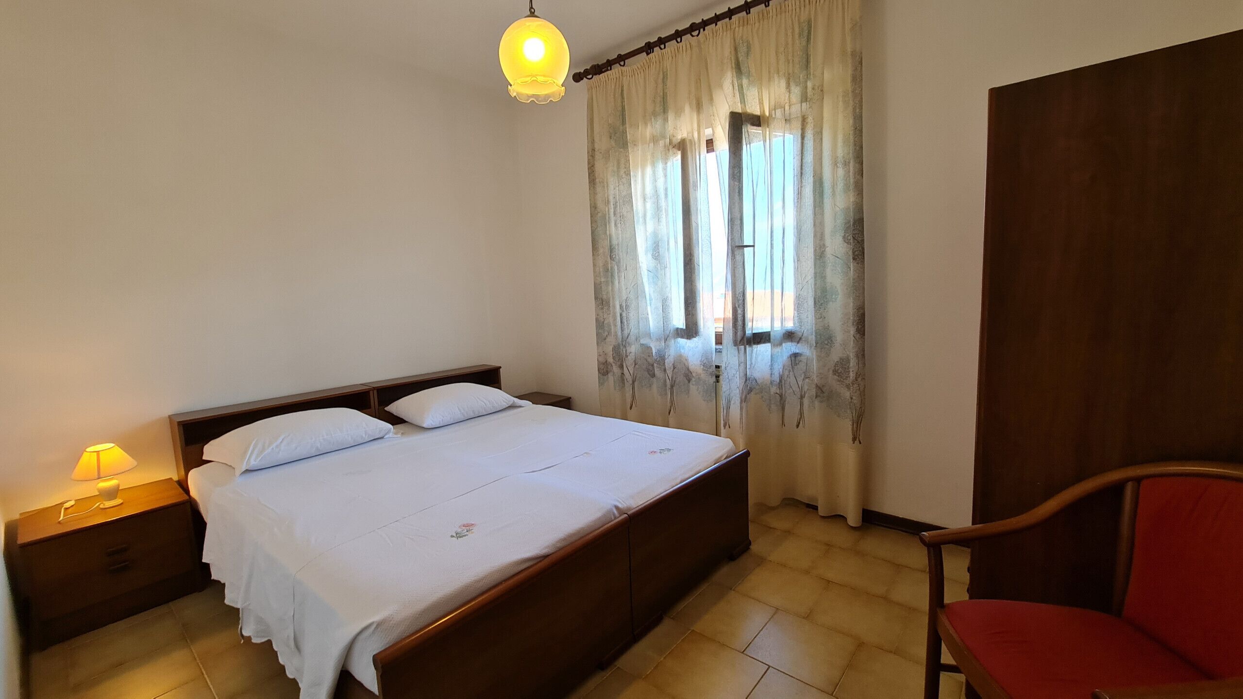Quarto com cama, mesa de cabeceira, luminária, cadeira e janela com cortinas.