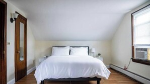Un dormitorio con cama, suelo de madera, mesita de noche, lámpara, ventana y puerta.