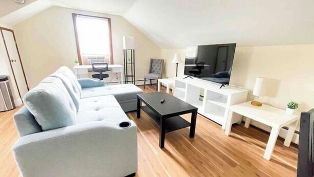 Una sala de estar moderna con un sofá seccional gris, una mesa de centro negra, un televisor sobre un mueble blanco y una pequeña mesa auxiliar blanca con una lámpara.