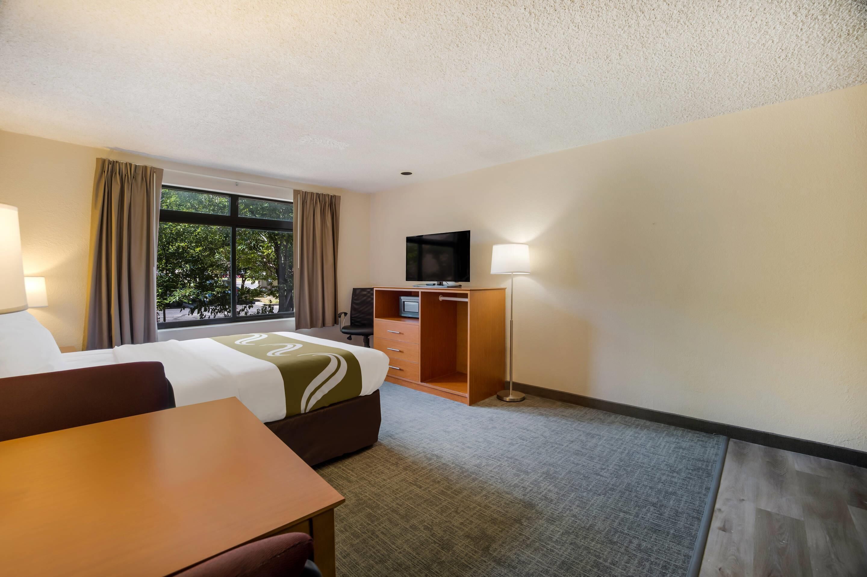 A hotel room with a bed, desk, TV, and a window with curtains.