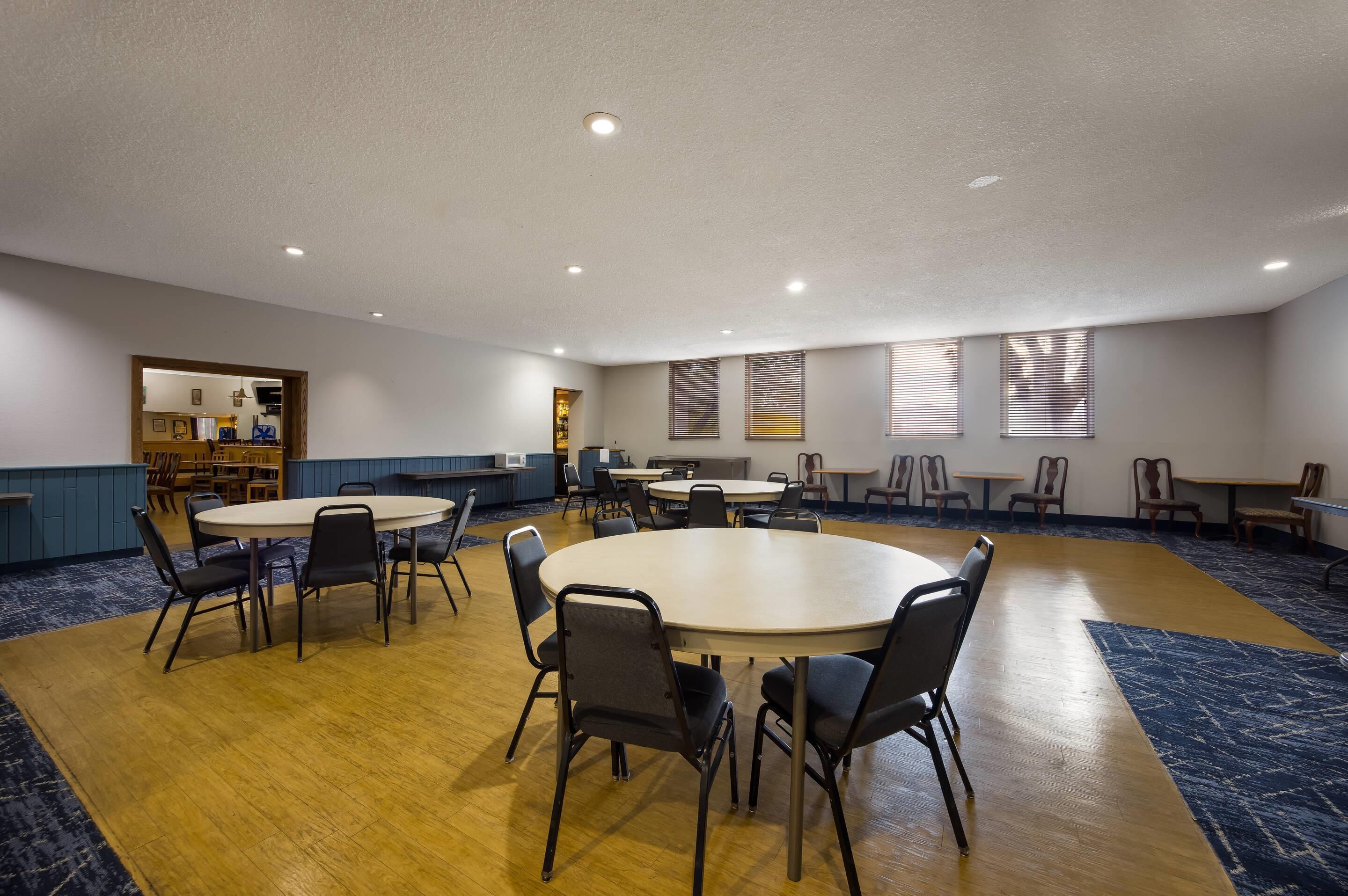 A spacious conference room with multiple tables and chairs, a kitchenette area, and large windows with blinds.