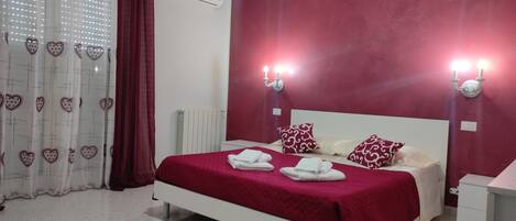 A neatly made bed with red bedding and white pillows, a white radiator, and a wall-mounted air conditioner.