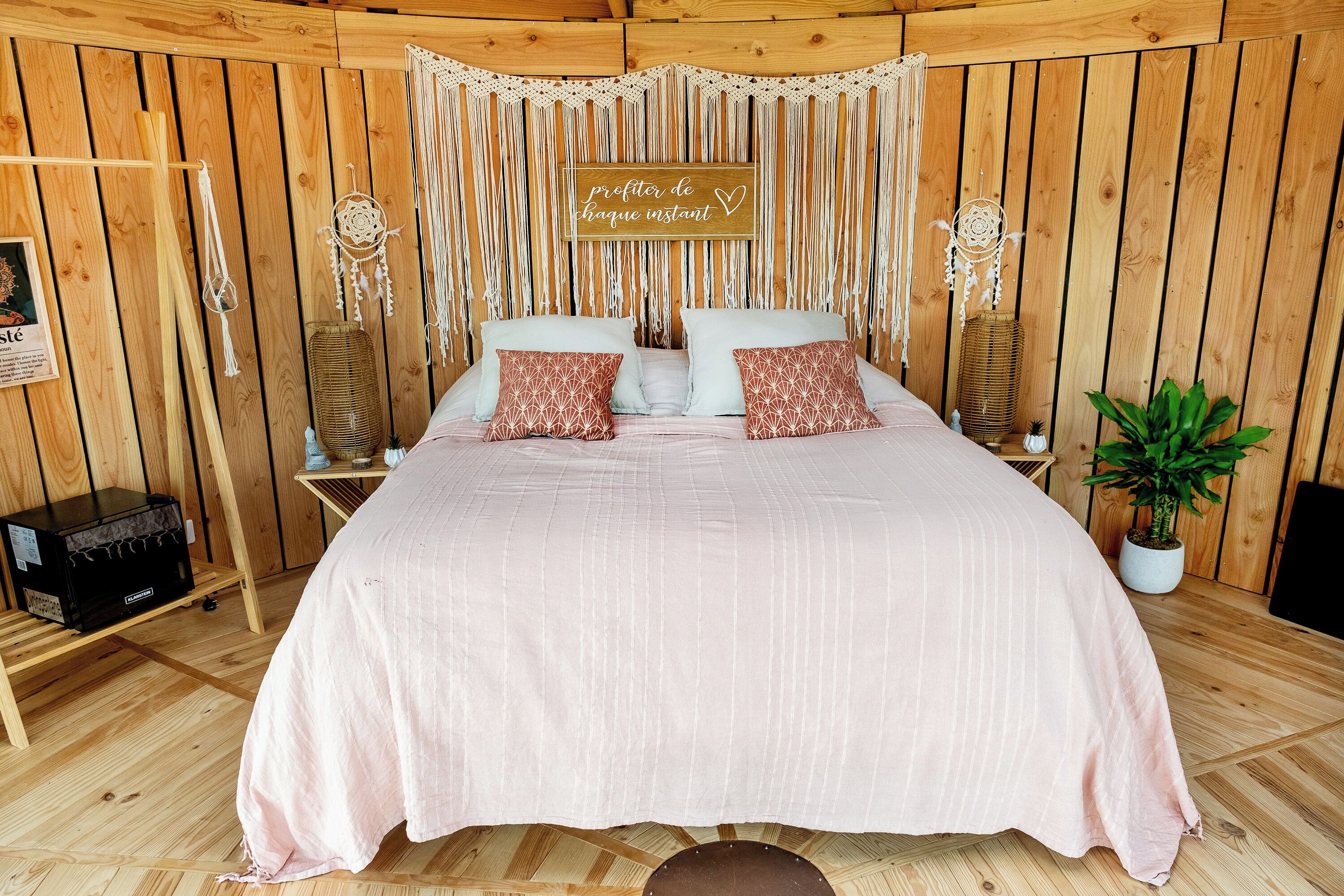 Een houten kamer met een bed, wanddecoratie van macramé en een bordje met de tekst 'profiter de chaque instant'.