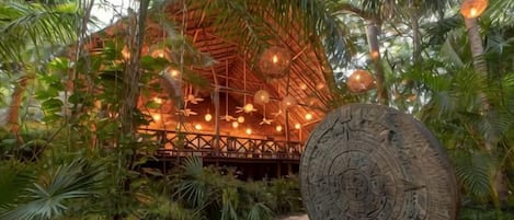 A rustic wooden structure with a thatched roof, surrounded by lush greenery and a stone pathway.