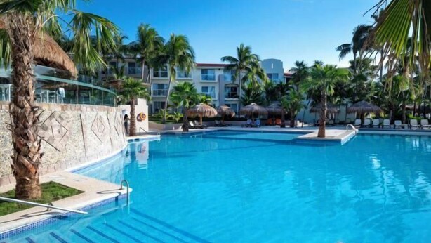 A clear blue swimming pool surrounded by palm trees and thatched umbrellas, with a view of hotel buildings in the background. - Viva Azteca by Wyndham, A Trademark All Inclusive Resort (PLAYA DEL CARMEN)