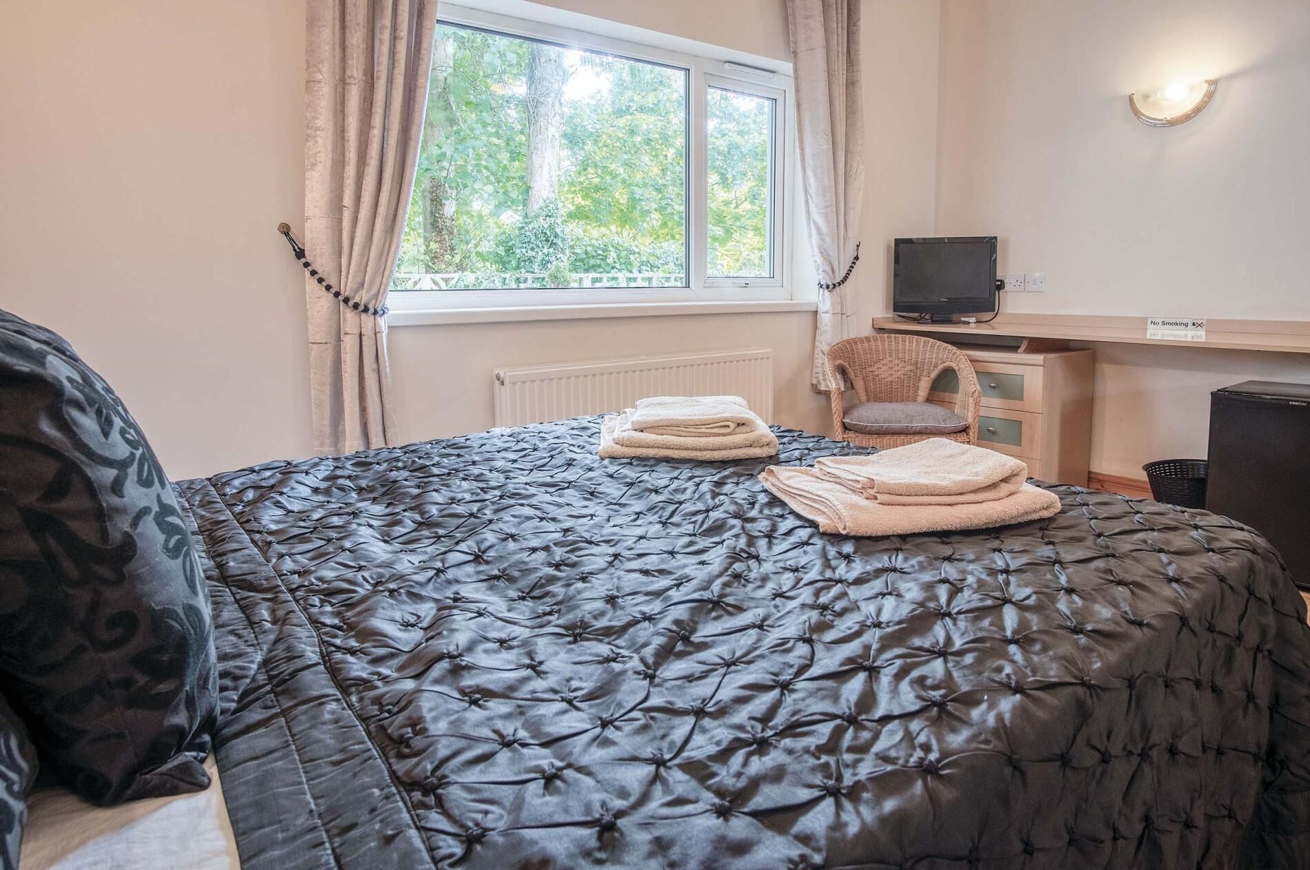 A neatly made bed with a dark quilt, two folded towels, a chair, a desk with a computer, and a window with curtains.