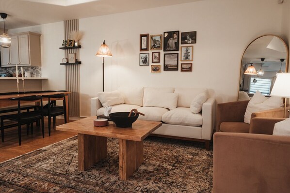 Un salon moderne avec un canapé, une table basse et un coin repas avec des chaises. On aperçoit au fond une cuisine équipée de placards et d’un plan de travail.
