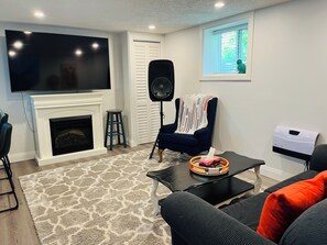 Un salon avec une cheminée, un téléviseur, un fauteuil confortable, une table basse et une fenêtre avec des stores.