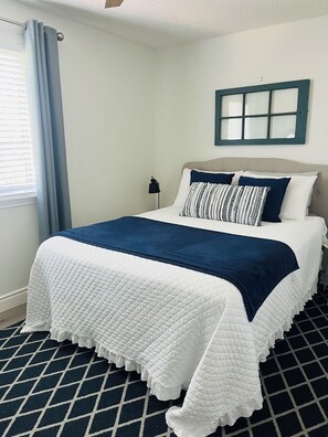 Un lit bien fait, recouvert d’une couette blanche et bleue, une lampe de chevet et une fenêtre avec des rideaux.