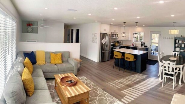 Una sala de estar moderna con un sofá gris, cojines amarillos, una mesa de centro de madera y un comedor con sillas blancas.