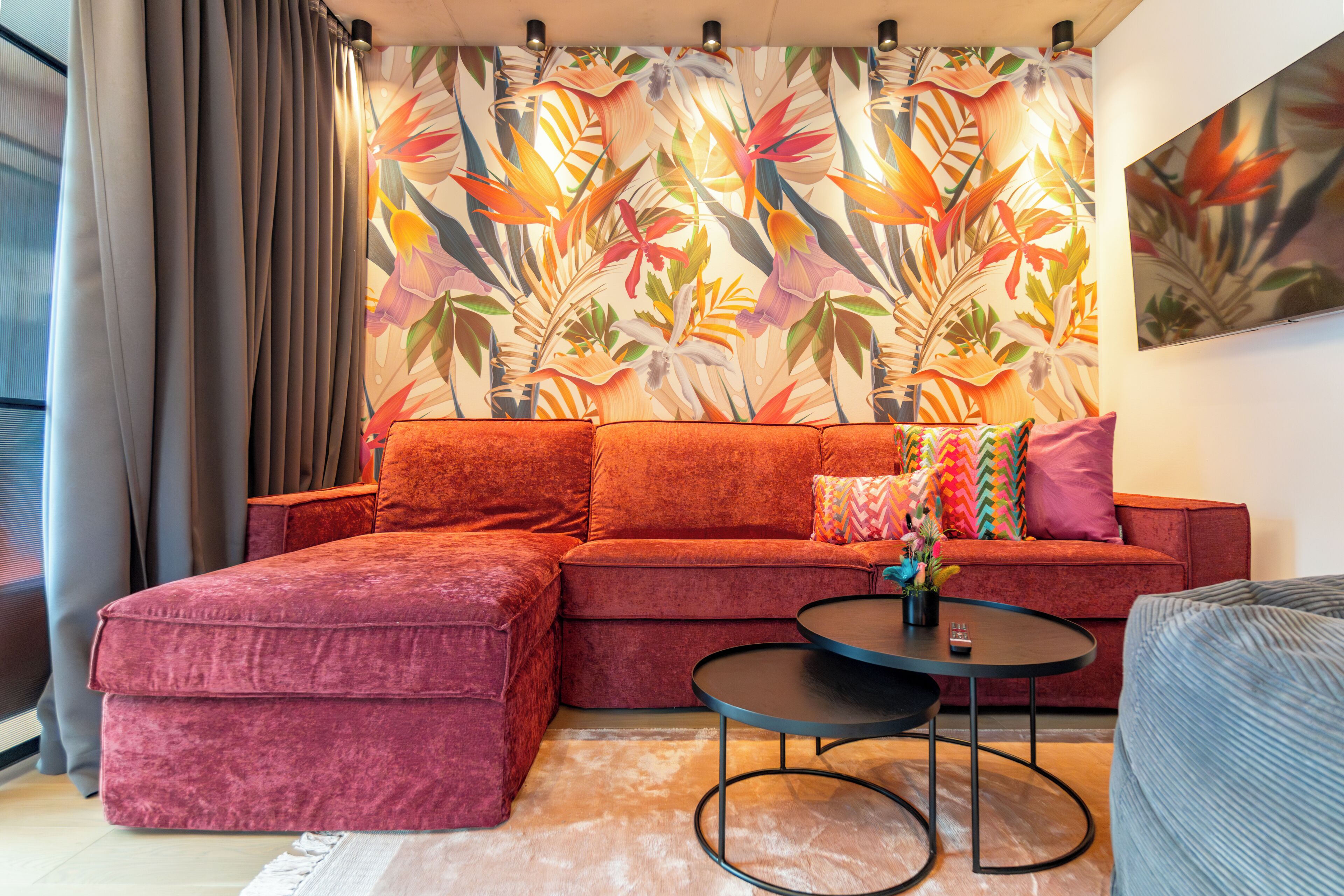 Un soggiorno moderno con un divano componibile rosso, un tavolino rotondo e una parete decorata con un vivace motivo a foglia tropicale.