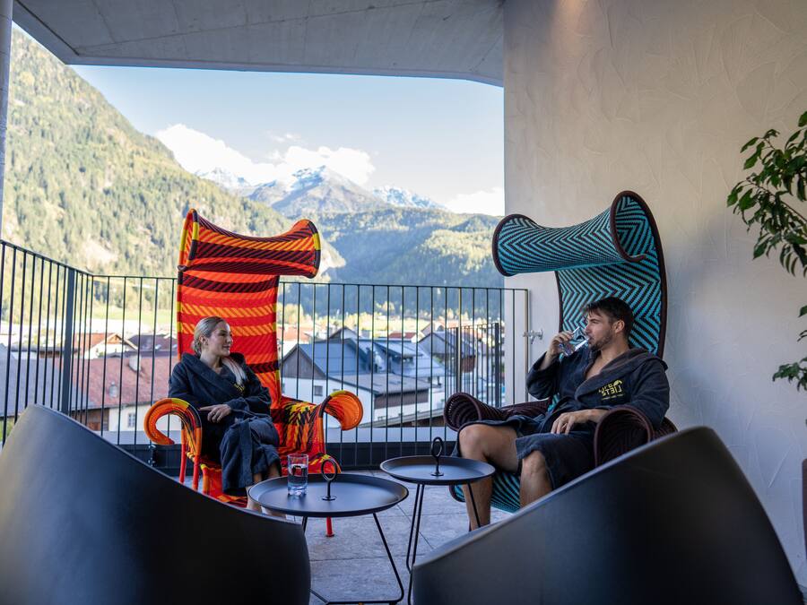 Dos personas sentadas en un balcón con vista a la montaña, una gran escultura abstracta y una barandilla.