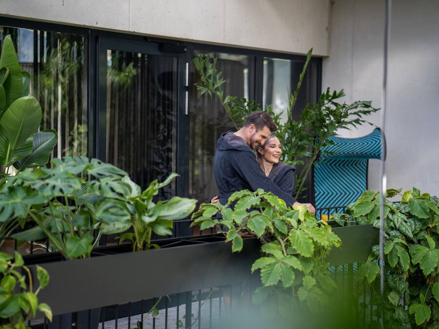 Un couple s’embrasse sur un balcon, avec une végétation luxuriante et un bâtiment moderne en arrière-plan.