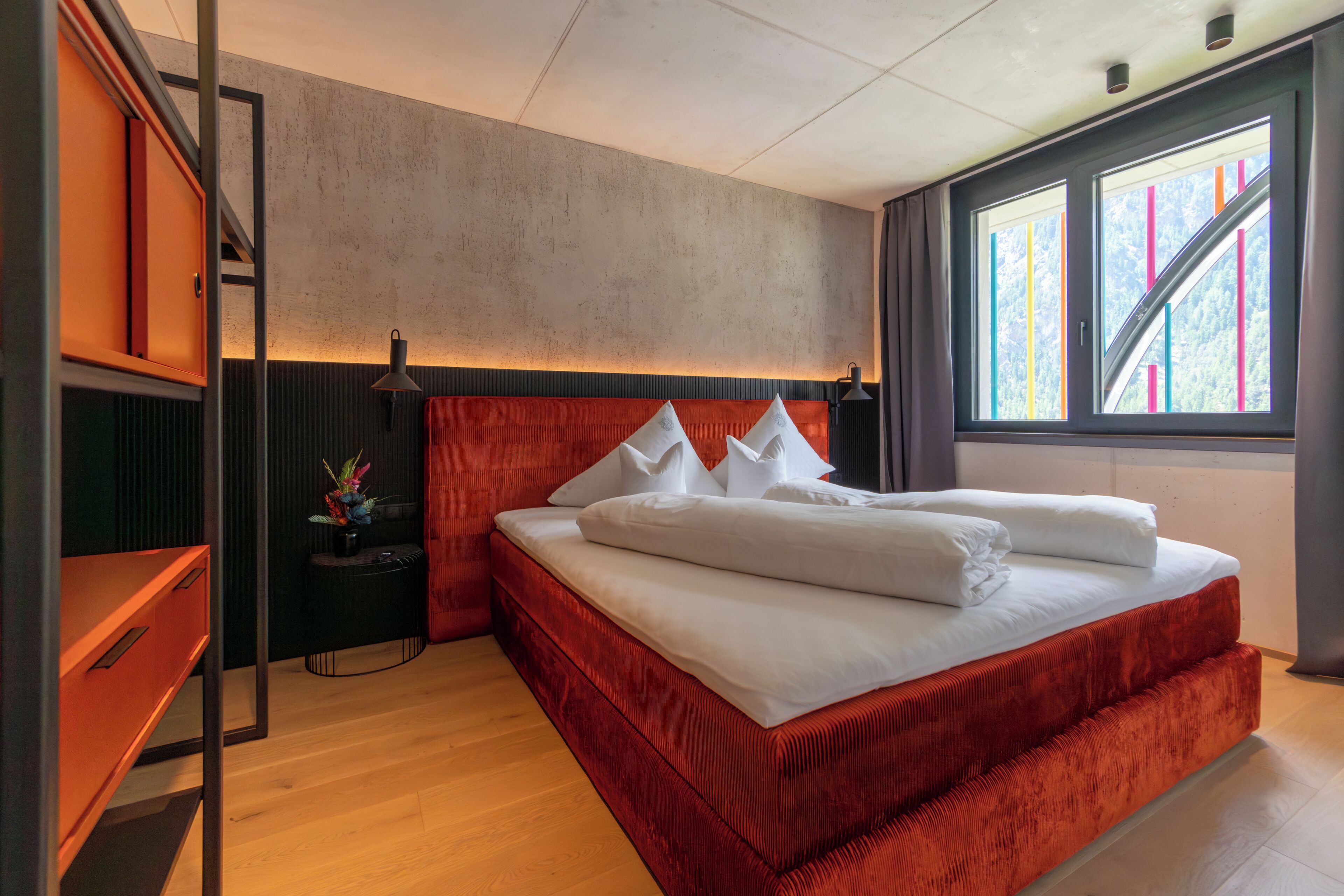 Une chambre moderne avec un grand lit, un bureau et une fenêtre donnant sur des bâtiments aux couleurs vives.