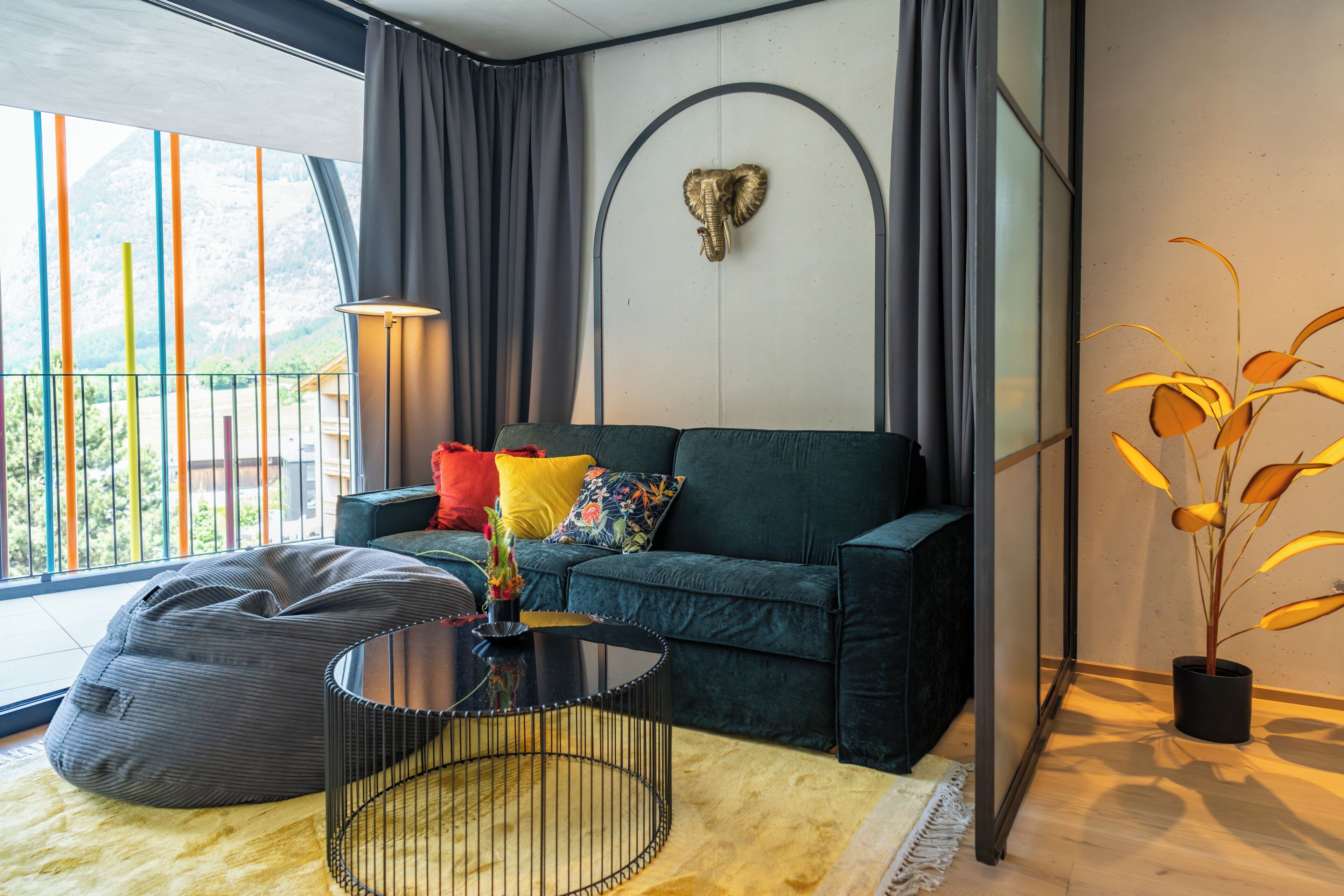 Ein modernes Wohnzimmer mit einem dunkelgrünen Sofa, bunten Kissen, einem schwarzen Couchtisch, einem grauen Sitzsack und einem dekorativen Elefantenkopf an der Wand.
