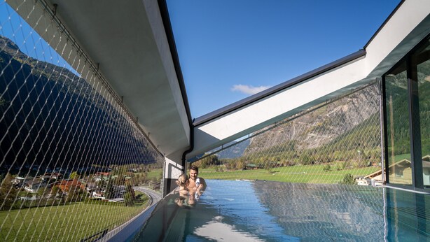 A glass-enclosed infinity pool with a mountain view, a clear sky, and a modern architectural design. - LIFESTEIL Appartementresort (Umhausen)