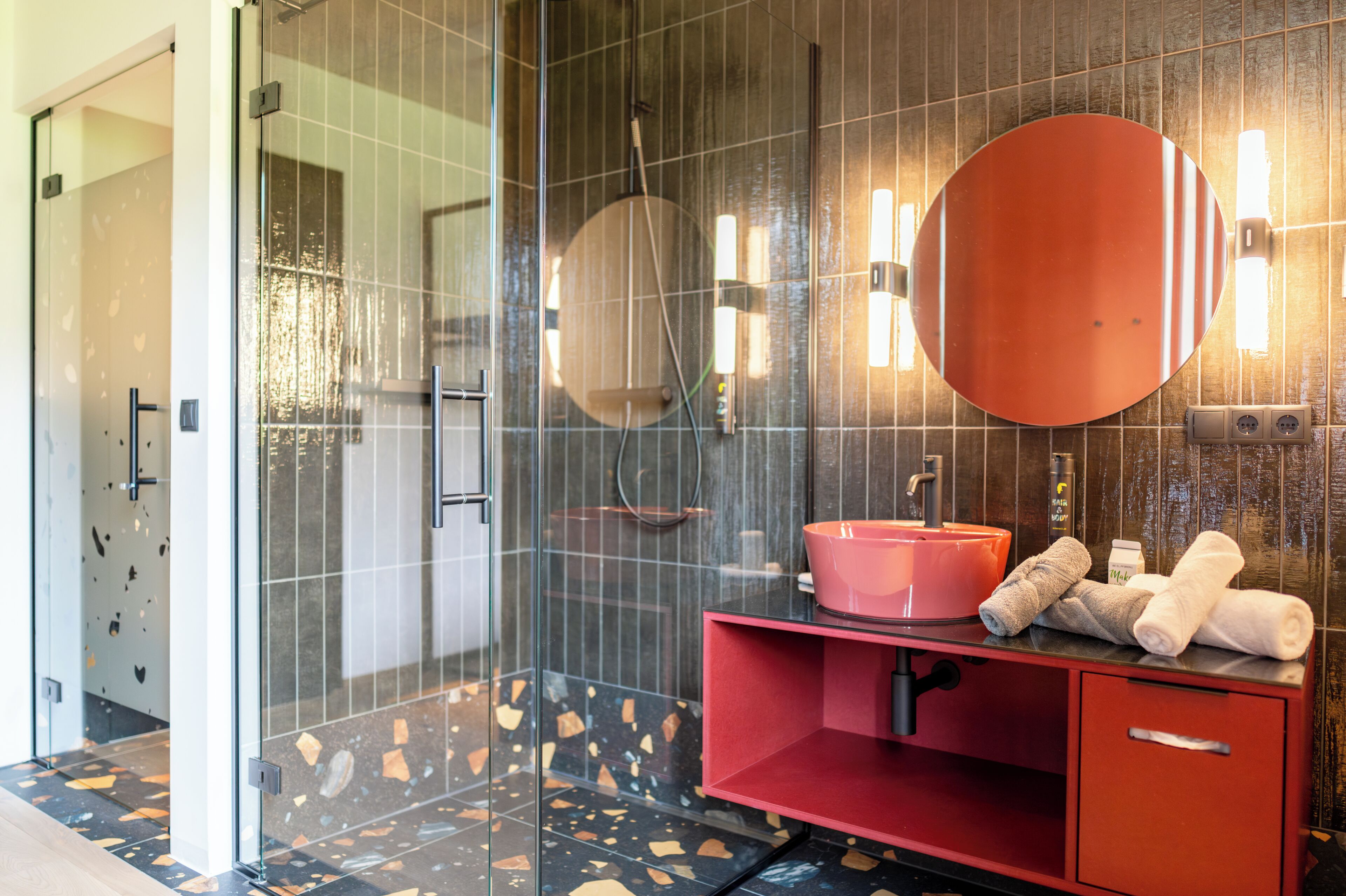 Een moderne badkamer met een douchecabine van glas, een rode wastafel met een roze wasbak en een grote spiegel.