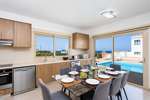 Une cuisine moderne avec des armoires en bois, un lave-vaisselle intégré et un coin repas avec une table dressée pour quatre personnes. De grandes fenêtres offrent une vue sur une piscine et un bâtiment situé de l’autre côté de l’eau.
