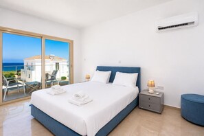 A hotel room with a bed, a nightstand, a lamp, a blue ottoman, and a balcony with a view of the sea.