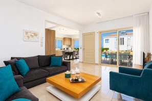 A modern living room with a sofa, coffee table, and teal chairs. There is a kitchen in the background and a view of the outdoors.