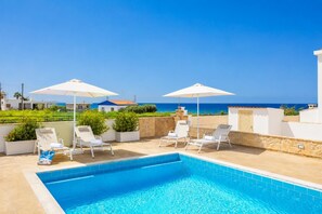 A pool area with lounge chairs and umbrellas, a clear blue sky, and a view of the sea.