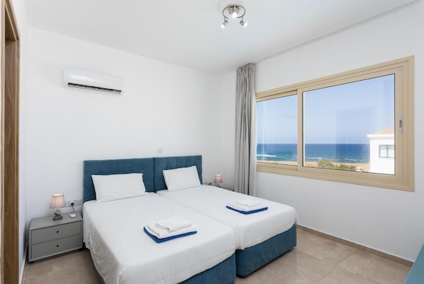A bedroom with two beds, a nightstand, an air conditioning unit, and a window with a view of the sea. - Villa Diana Seaside - Four Bedroom Villa, Sleeps 8 (Pomos)