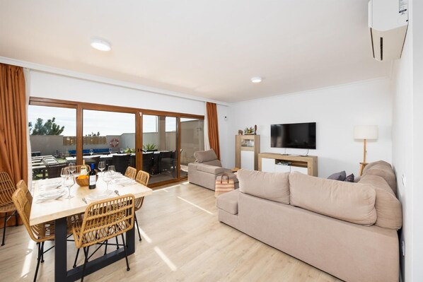 En moderne stue med sofa, fjernsyn og spiseområde med et bord dækket til fire personer.
