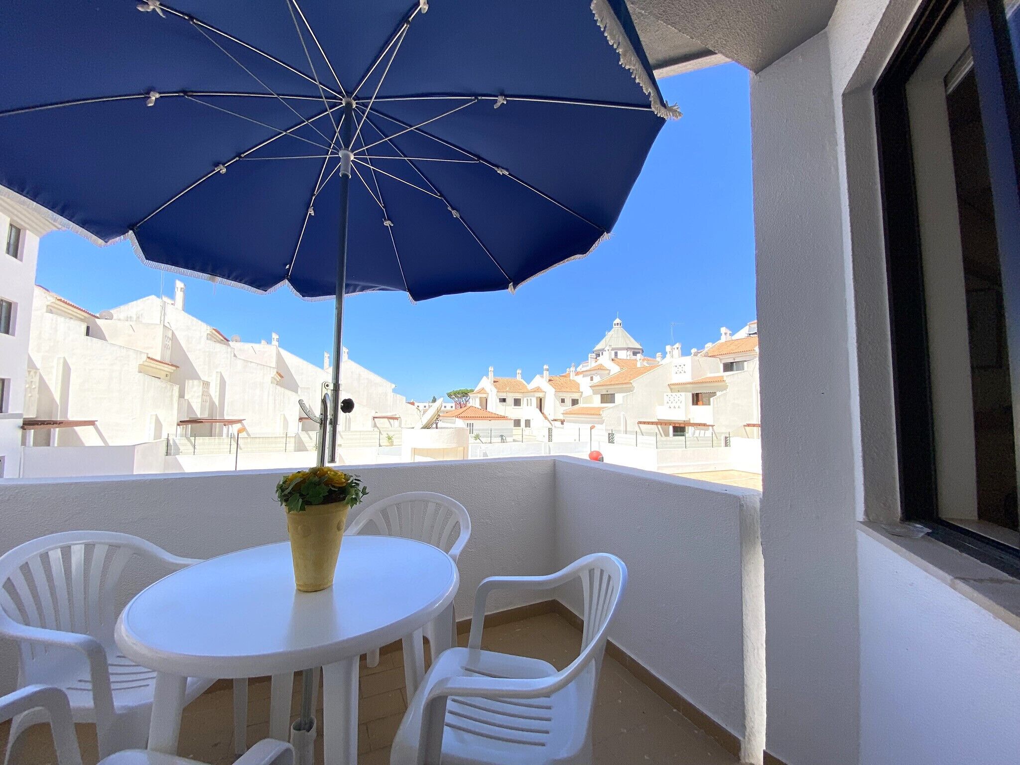 Een balkon met een blauwe parasol, witte stoelen en een kleine tafel met een bloempot.