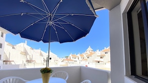 Un balcon avec un parasol bleu, des chaises blanches et une petite table sur laquelle est posée une plante en pot.