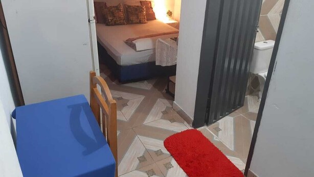 Een smalle gang die leidt naar een slaapkamer met een bed, een nachtkastje en een bijzettafeltje. Een rood tapijt ligt op de vloer, en een blauwe stoel is in de voorgrond zichtbaar.
