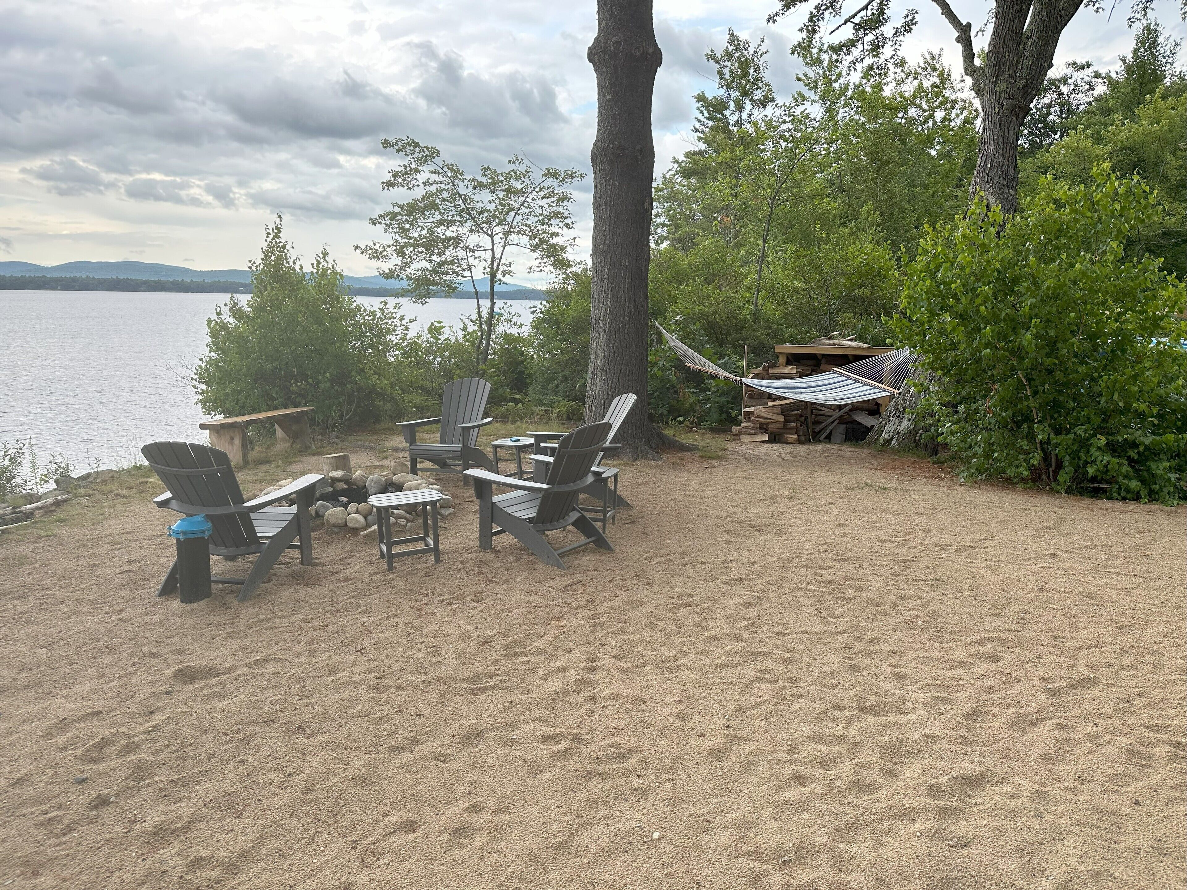 A lakeside outdoor seating area with Adirondack chairs, a fire pit, and a hammock.