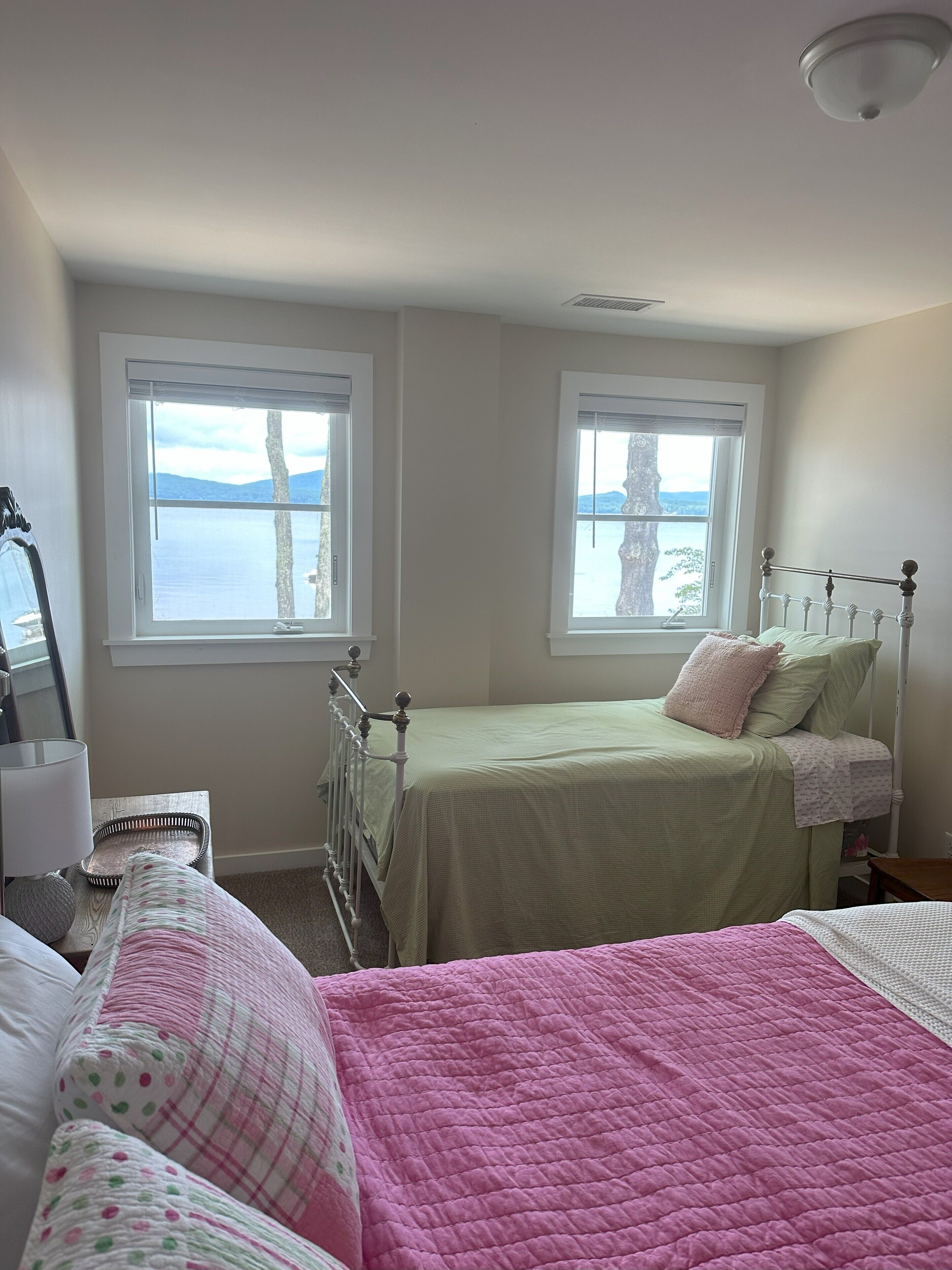 Une chambre avec deux lits, de grandes fenêtres donnant sur la mer, un ventilateur de plafond et une table de chevet avec une lampe.