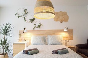 A well-lit bedroom with a wooden headboard, two bedside tables, a large bed with rolled towels, and decorative wall art.
