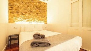 Une chambre d’hôtel avec un mur en pierre, un lit avec du linge de maison blanc, une petite table de chevet et une fenêtre avec des stores.