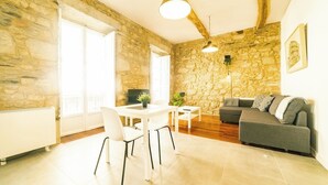 Un salon aux murs en pierre, une table blanche avec des chaises, un canapé gris et un luminaire suspendu.