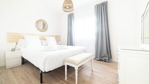 A bedroom with a wooden headboard, a white bed with two pillows, a bedside table with a lamp, a bench, and a large window with grey curtains. - House O Carteiro "Cedeira". (Cedeira)